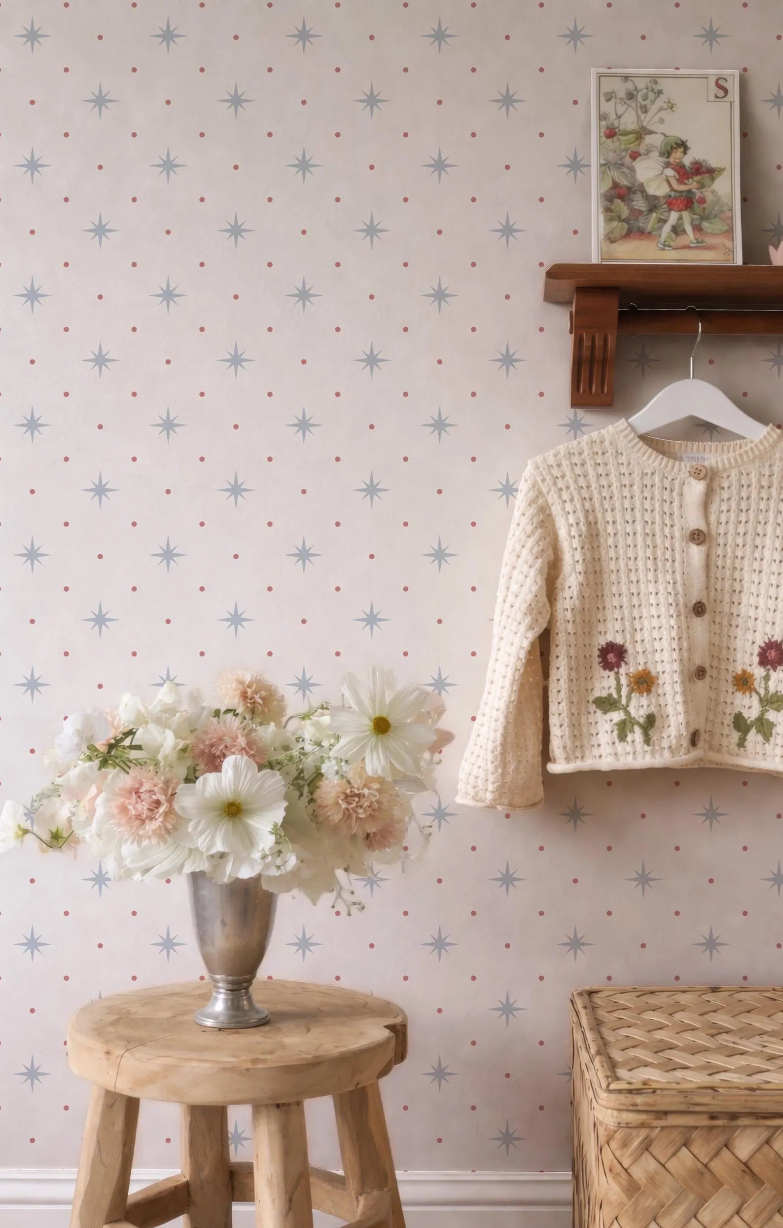 a cozy and charming interior scene with a wooden stool holding a vase filled with white and pink flowers, a wooden shelf displaying a white sweater with floral embroidery, and a wooden shelf with a framed picture. The wall behind these items is decorated with a pattern of blue and white stars.