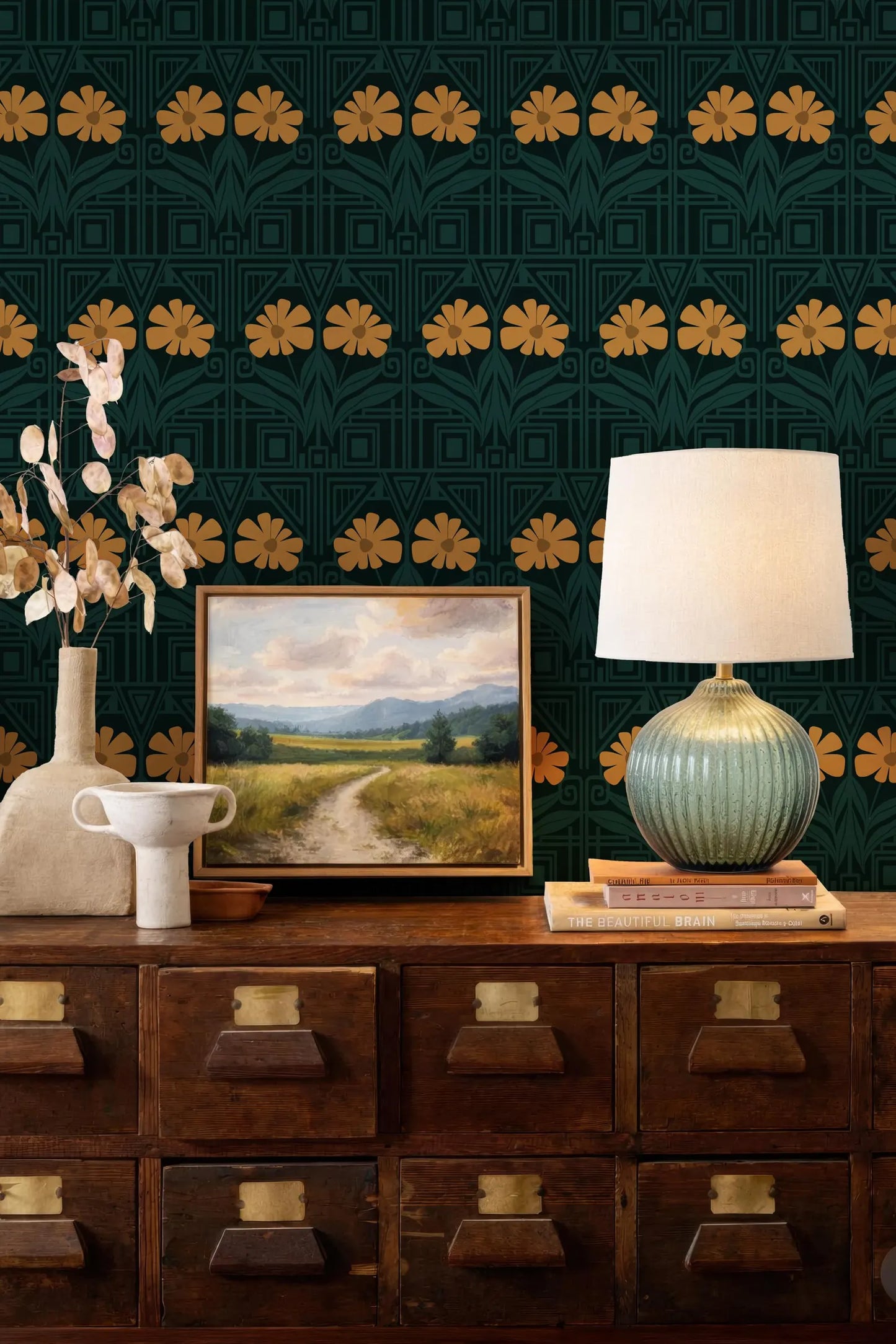 a wooden dresser with a painting, a lamp, and a vase on top. The dresser has multiple drawers and a cabinet below it. The wall behind the dresser has a pattern of yellow flowers.