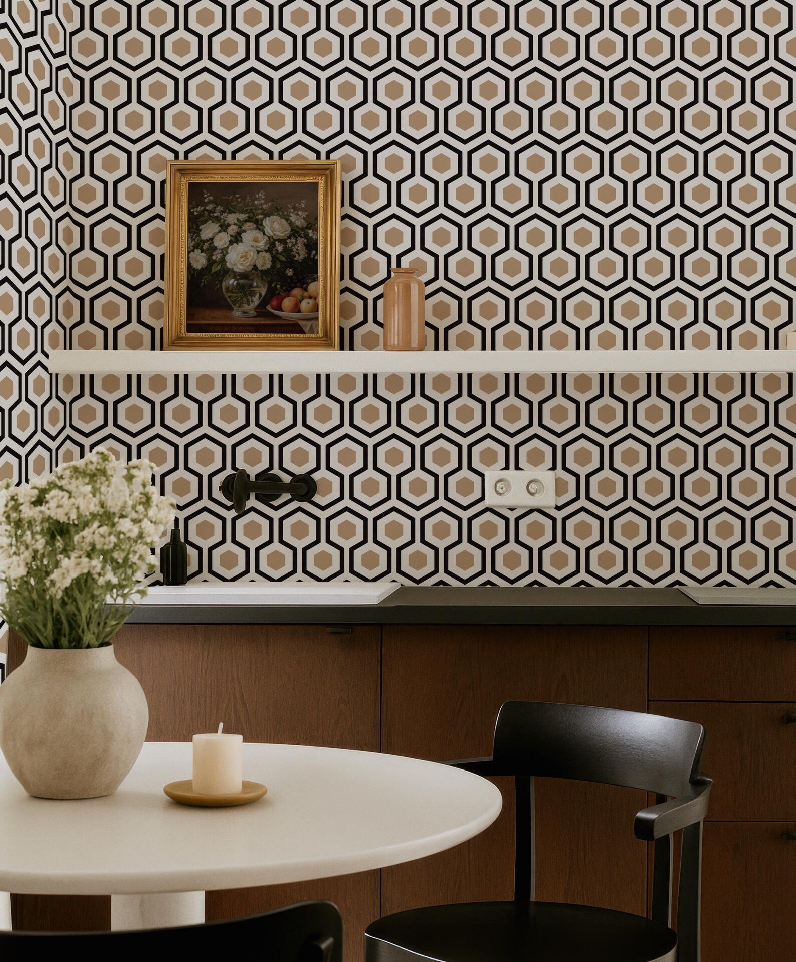 a modern kitchen with a white table, a vase of white flowers, and a framed picture on the wall. The kitchen has a geometric patterned wallpaper, and there is a shelf with a vase and a candle on it.