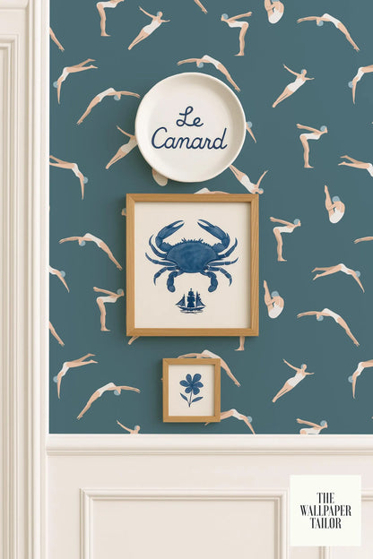 a wallpapered wall with a framed picture of a crab, a framed picture of a flower, and a framed picture of a person diving. The wallpapered wall has a pattern of swimming figures, and there is a plate with the word "Camard" written on it.