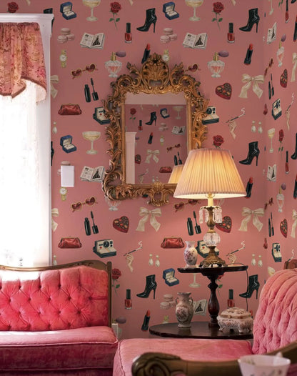 a cozy and stylishly decorated living room with a pink color scheme. The room features a pink sofa, a pink armchair, and a pink ottoman. The walls are adorned with a pink wallpaper that showcases various fashion and beauty-related icons, including high heels, makeup, and accessories. A gold-framed mirror is mounted on the wall, and a lamp with a beige shade is placed on a small table in front of the sofa.
