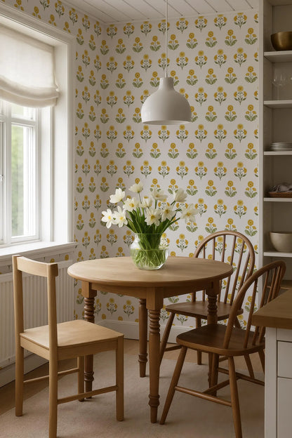 a cozy dining area with a round wooden table, two wooden chairs, and a vase of white flowers on the table. The walls are covered in a patterned wallpaper with yellow and green floral designs. A window is visible on the left side of the image, allowing natural light to enter the space.