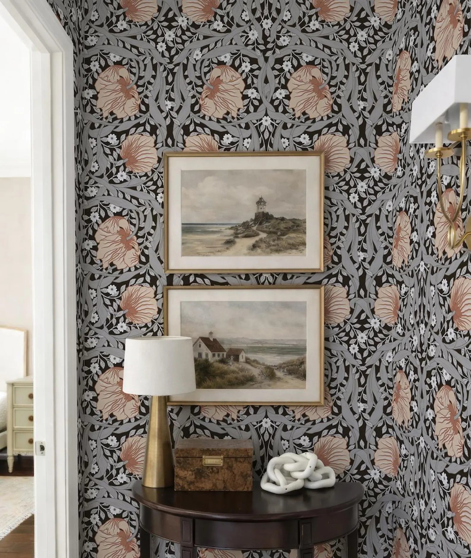 a corner of a room with a dark wood table, a lamp, and two framed paintings hanging on the wall above it. The wall is covered in a floral patterned wallpaper, and the overall color scheme is a combination of black, white, and orange.