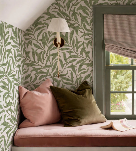 a cozy corner of a room with a pink and green patterned wallpaper, a green and brown throw pillow, and a lamp with a white shade. There is a window with a green frame and a white curtain, and a book is placed on the windowsill.