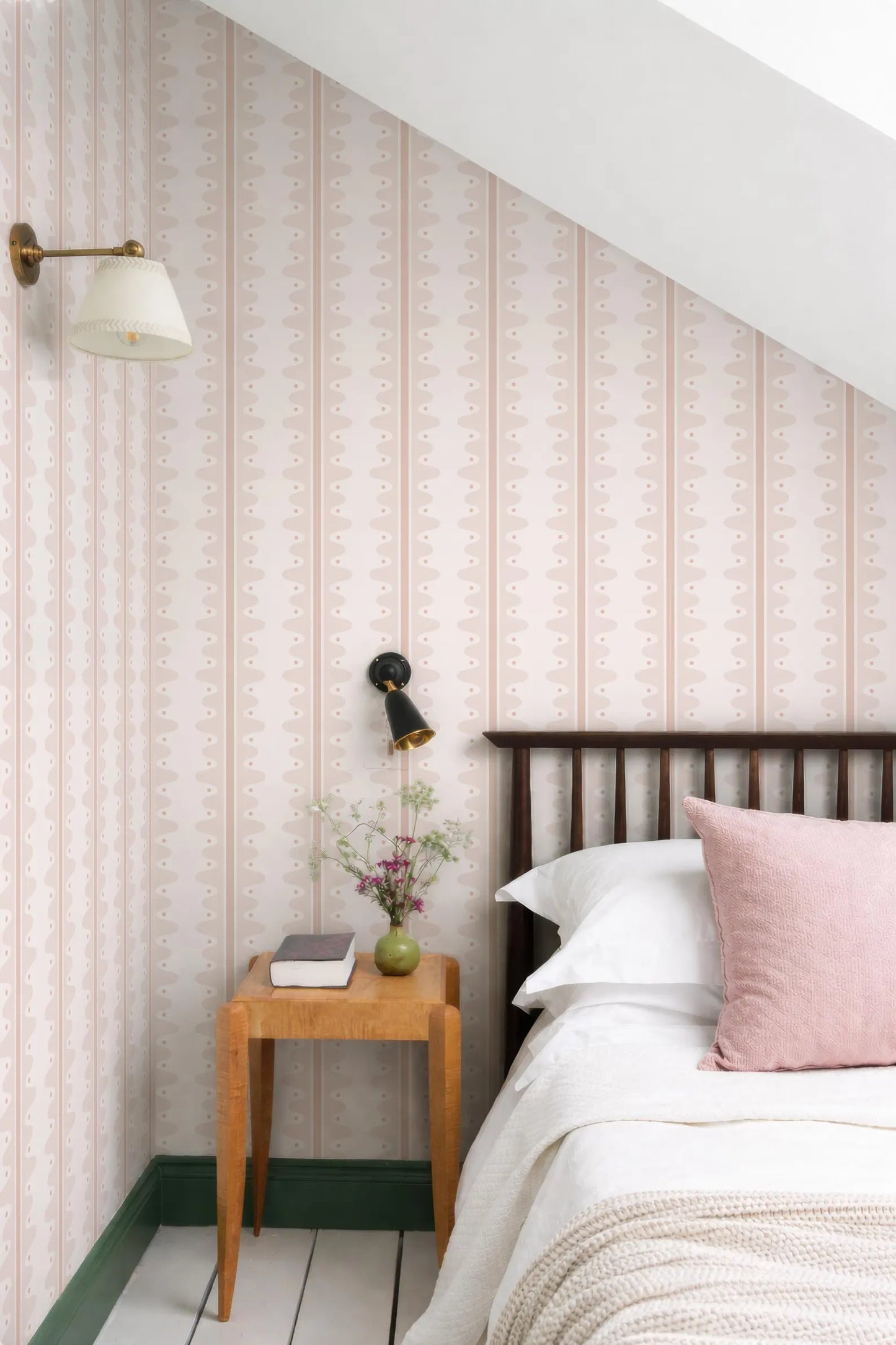 a cozy bedroom with a wooden bed frame, a wooden nightstand, and a lamp. The walls are covered in pink and white striped wallpaper, and there is a vase of flowers on the nightstand.