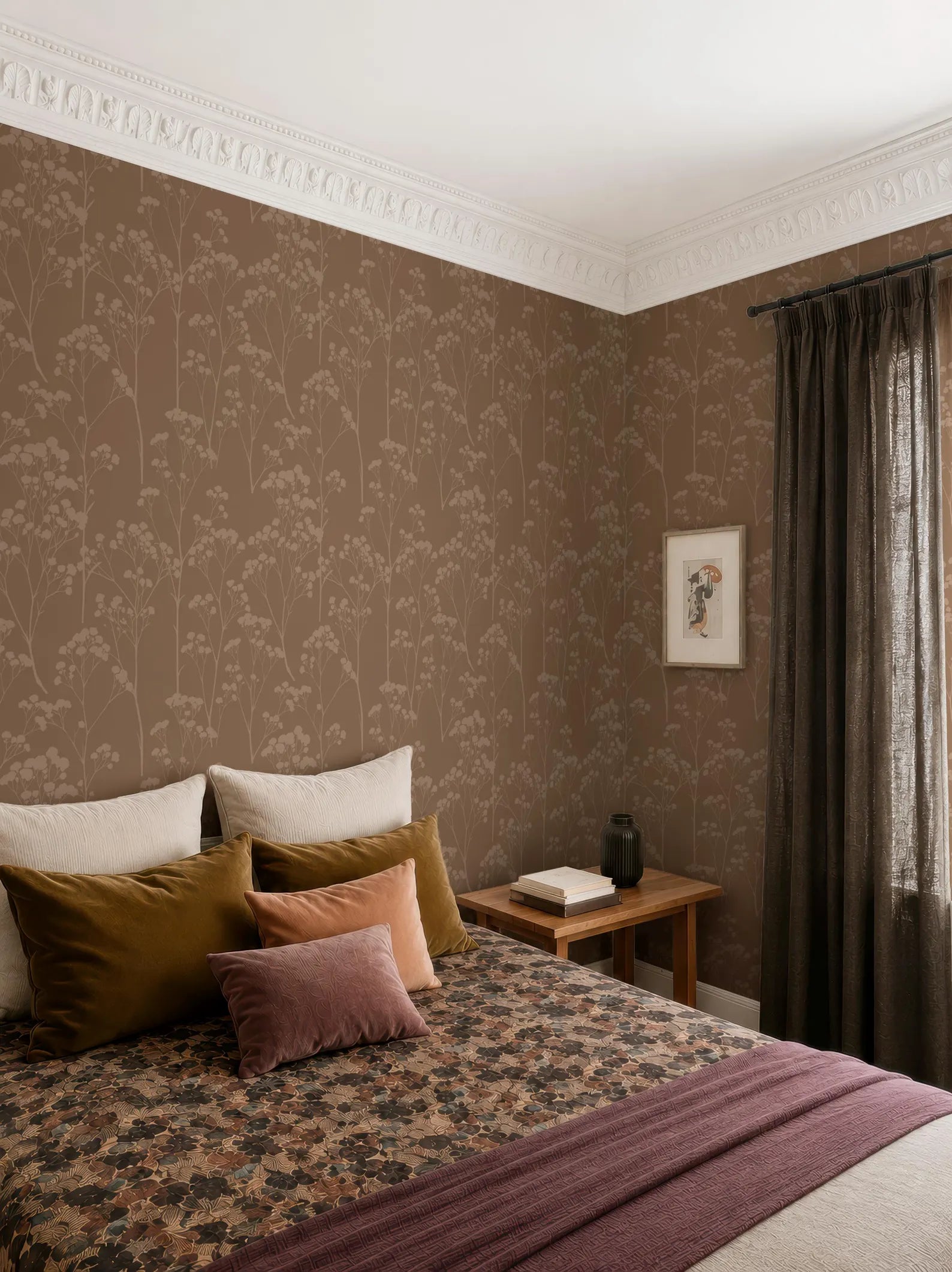 a cozy bedroom with a large bed adorned with a floral bedspread and a variety of colorful pillows. The room features a wooden nightstand with a book and a vase, and a window with curtains. The walls are covered in a patterned wallpaper, and the overall color scheme is warm and inviting.
