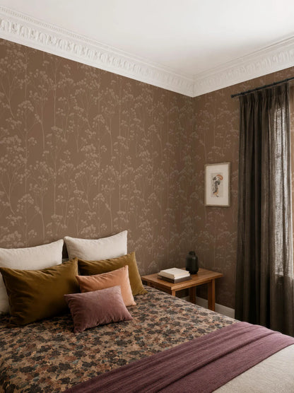 a cozy bedroom with a large bed adorned with a floral bedspread and a variety of colorful pillows. The room features a wooden nightstand with a book and a vase, and a window with curtains. The walls are covered in a patterned wallpaper, and the overall color scheme is warm and inviting.
