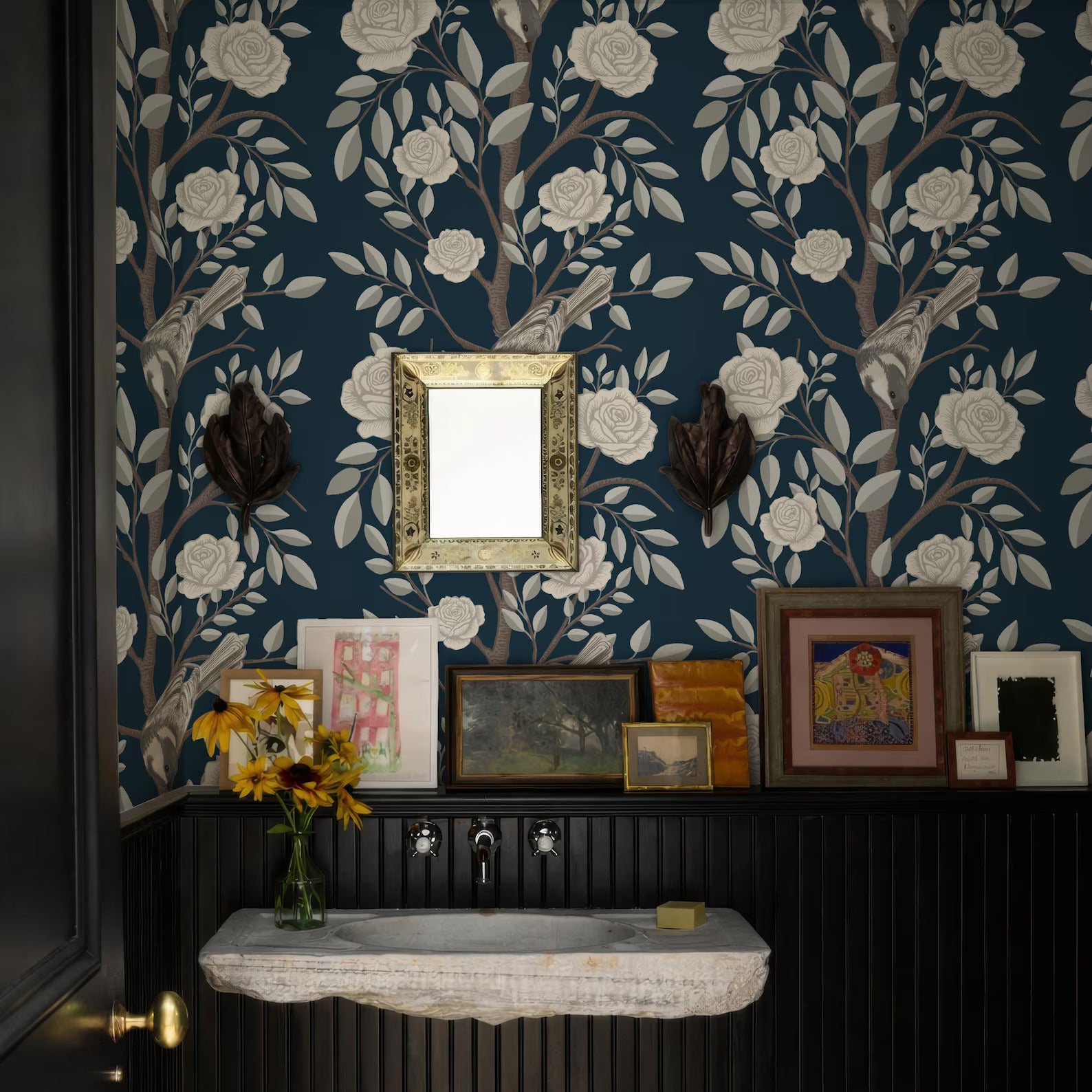 a bathroom with a blue floral wallpaper, a white sink, and a wooden shelf with framed pictures and a vase of yellow flowers.