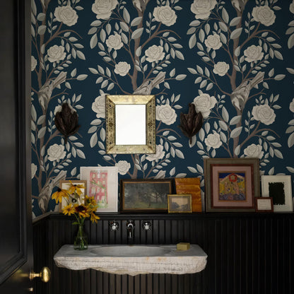 a bathroom with a blue floral wallpaper, a white sink, and a wooden shelf with framed pictures and a vase of yellow flowers.