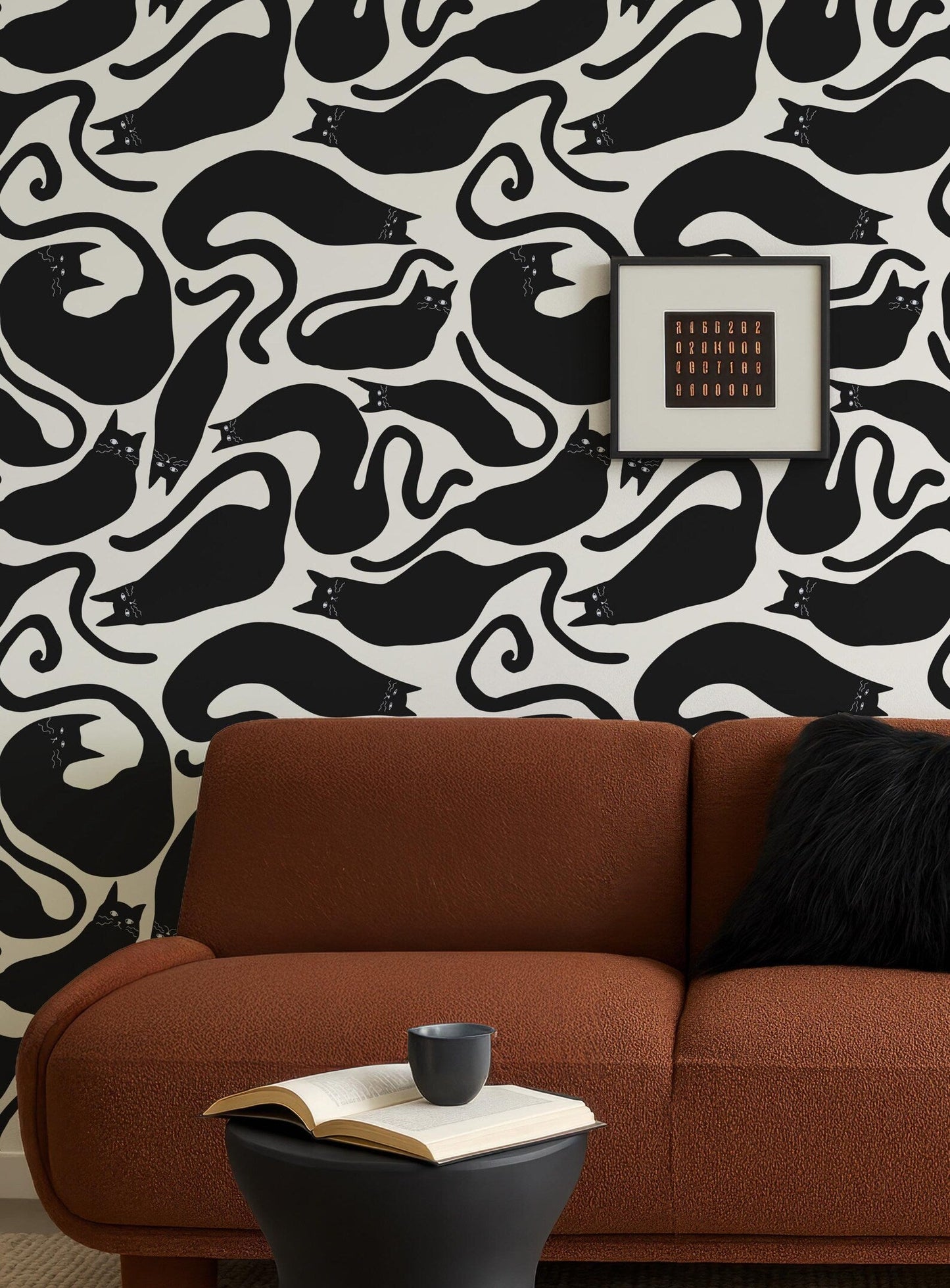 a cozy living room with a brown sofa, a black and white cat-themed wallpaper, and a framed picture on the wall. On the sofa, there is a black and white cat plushie and a black and white cat-themed coffee mug. In front of the sofa, there is an open book and a small black side table.