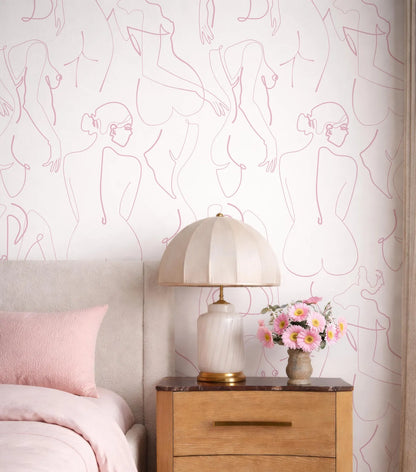 a bedroom setting with a bed, a wooden nightstand, and a lamp with a white shade. The wall behind the nightstand has a repeating pattern of pink and white abstract human figures, creating a visually striking and artistic backdrop.