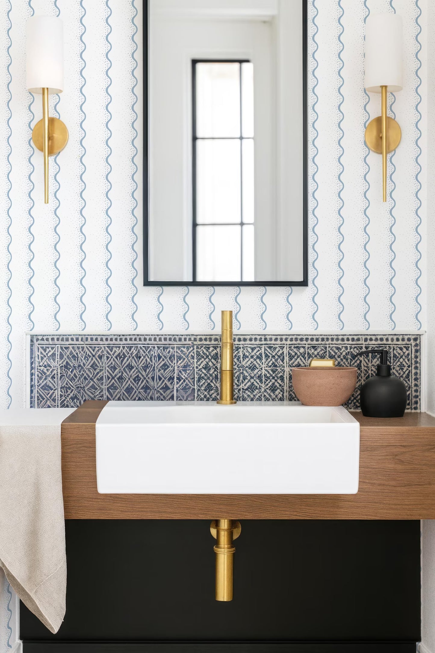 a modern bathroom with a white sink, a gold faucet, and a black countertop. The walls are adorned with blue and white patterned wallpaper, and there are two gold wall sconces on either side of the mirror.
