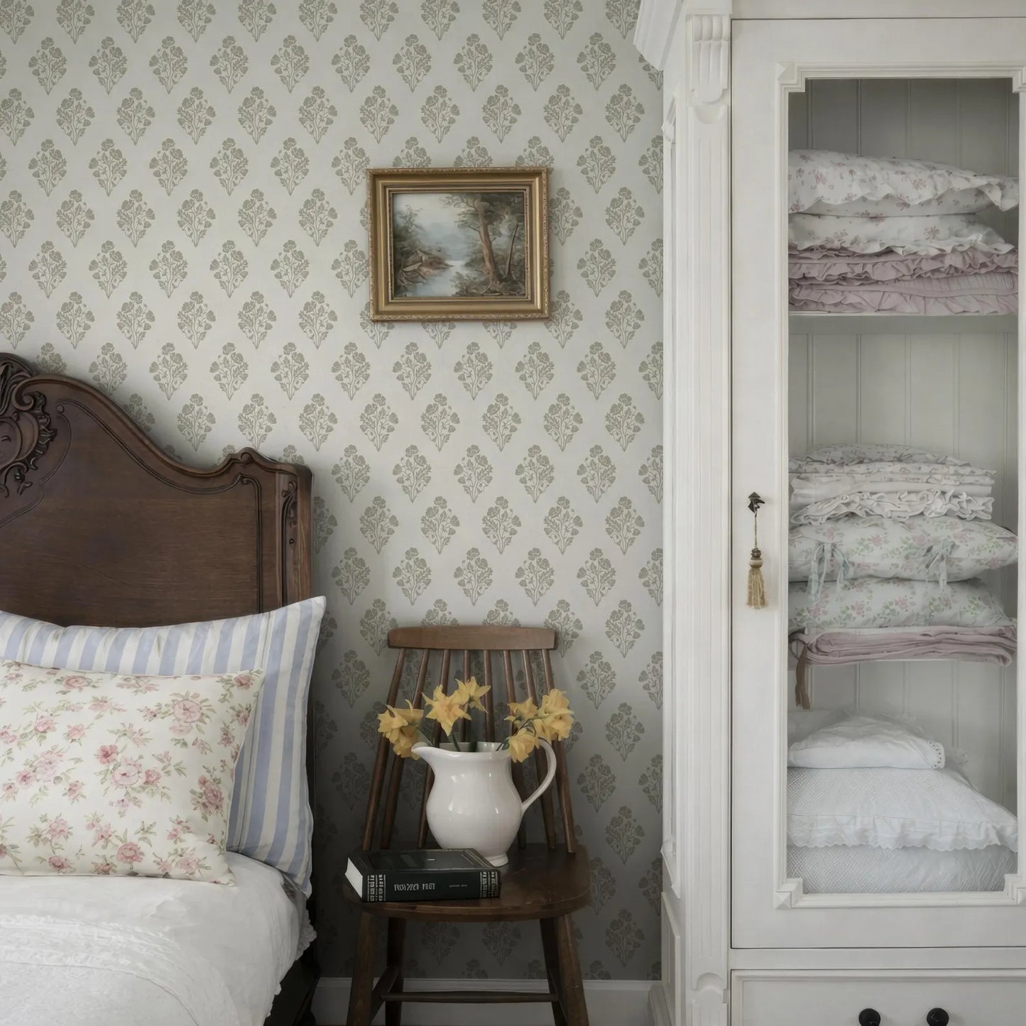 a cozy bedroom scene with a wooden headboard, a white wall with a floral wallpaper, a wooden chair with a vase of yellow flowers, and a white wooden cabinet with folded blankets.