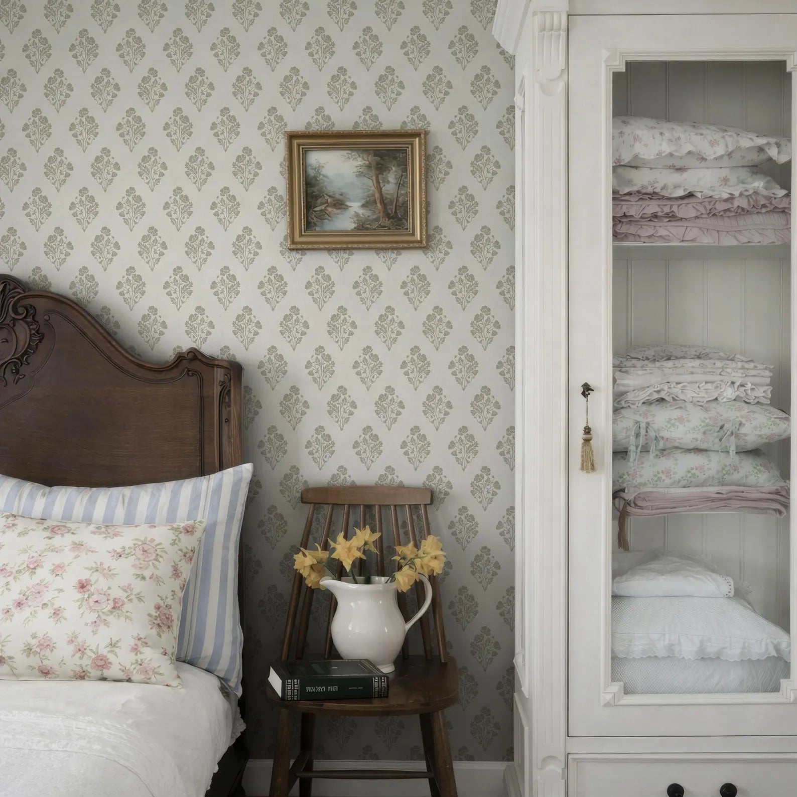 a cozy bedroom scene with a wooden headboard, a white wall with a floral wallpaper, a wooden chair with a vase of yellow flowers, and a white wooden cabinet with folded blankets.