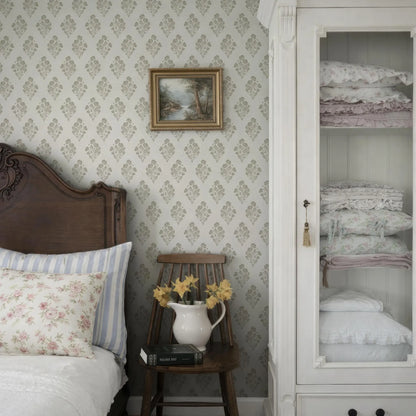 a cozy bedroom scene with a wooden headboard, a white wall with a floral wallpaper, a wooden chair with a vase of yellow flowers, and a white wooden cabinet with folded blankets.