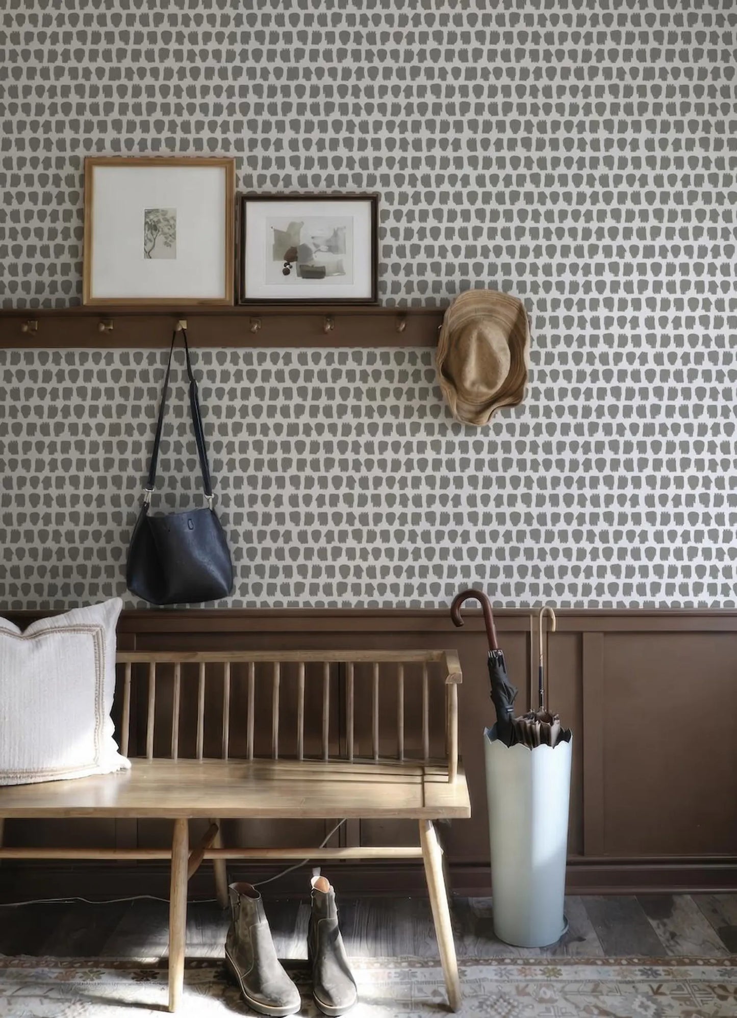 a cozy entryway with a wooden bench, a wall with a patterned wallpaper, and a shelf holding a hat, umbrella, and handbag.