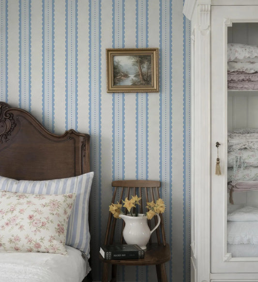 a cozy bedroom scene with a wooden headboard, a white and blue striped wallpaper, and a wooden chair with a vase of yellow flowers.