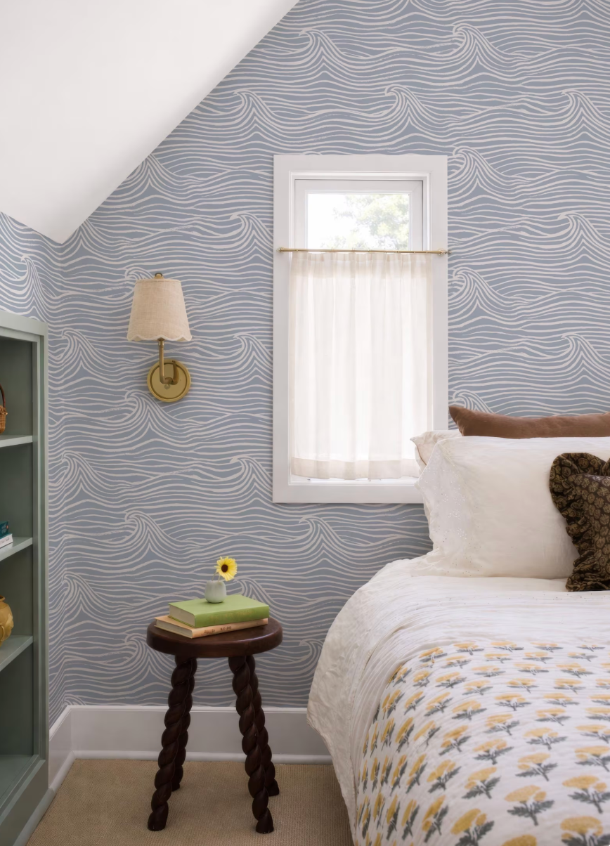 a cozy bedroom with a bed, a small wooden stool, and a window with a white curtain. The walls are covered in a blue and white wave wallpaper, and there is a small table with a vase of flowers and a book on it.