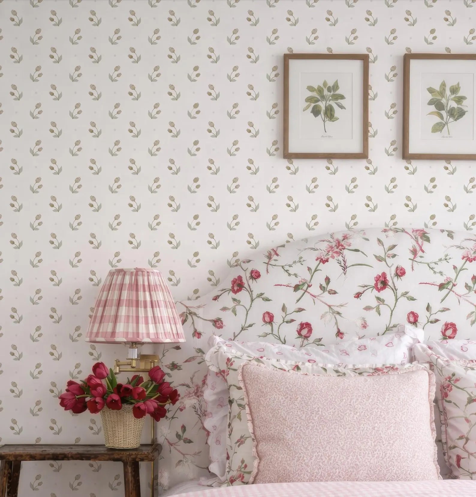 a bedroom with a floral print wallpaper, a pink and white striped lamp, a wooden nightstand with a vase of red flowers, and two framed botanical prints on the wall.