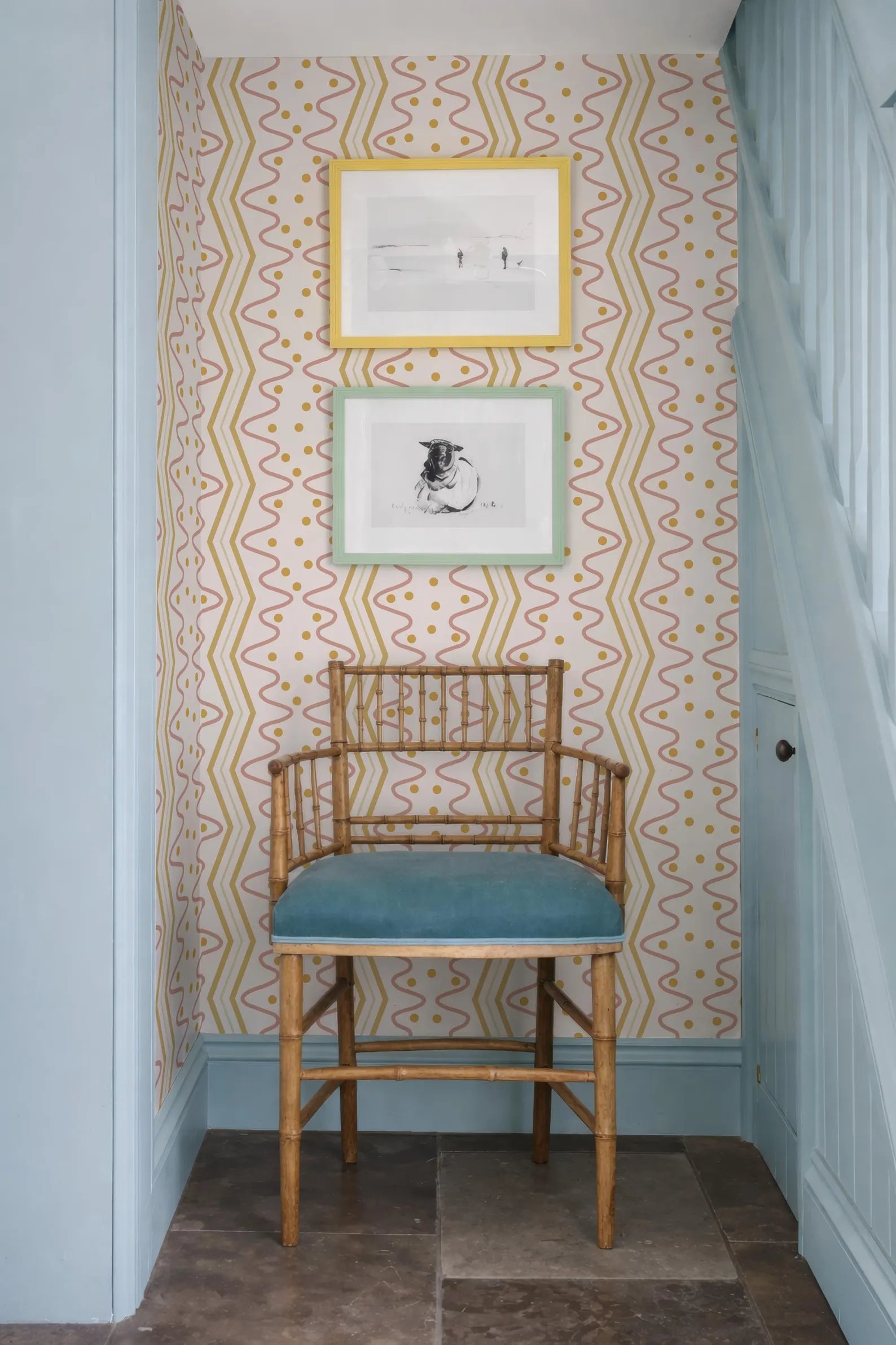 a wooden chair with a blue cushion, positioned in front of a wall with a patterned wallpaper. Above the chair, there are two framed pictures hanging on the wall.