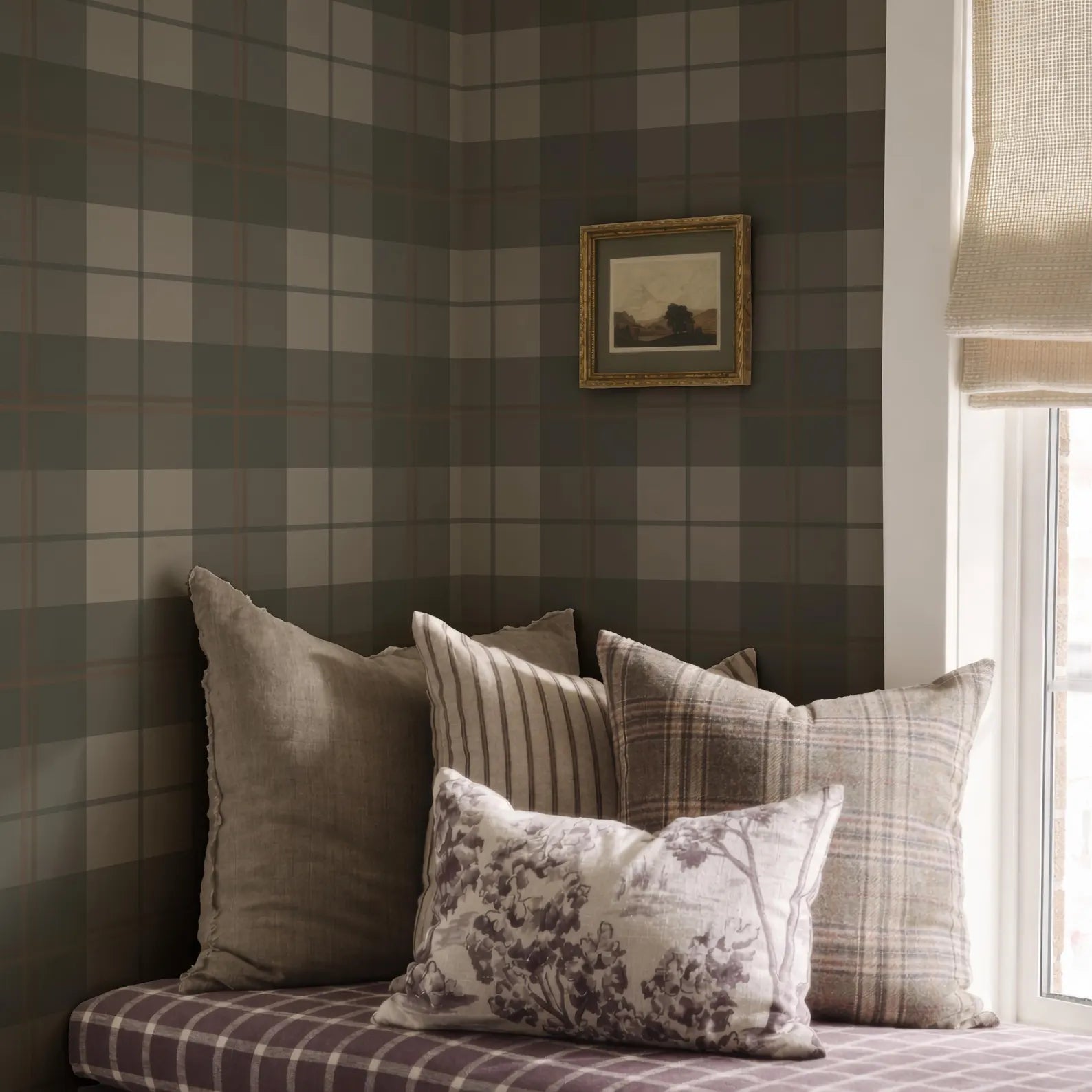 a cozy corner of a room with a plaid pattern on the wall and a window with a white curtain. The room has a mix of neutral and patterned pillows on a bed, creating a warm and inviting atmosphere.