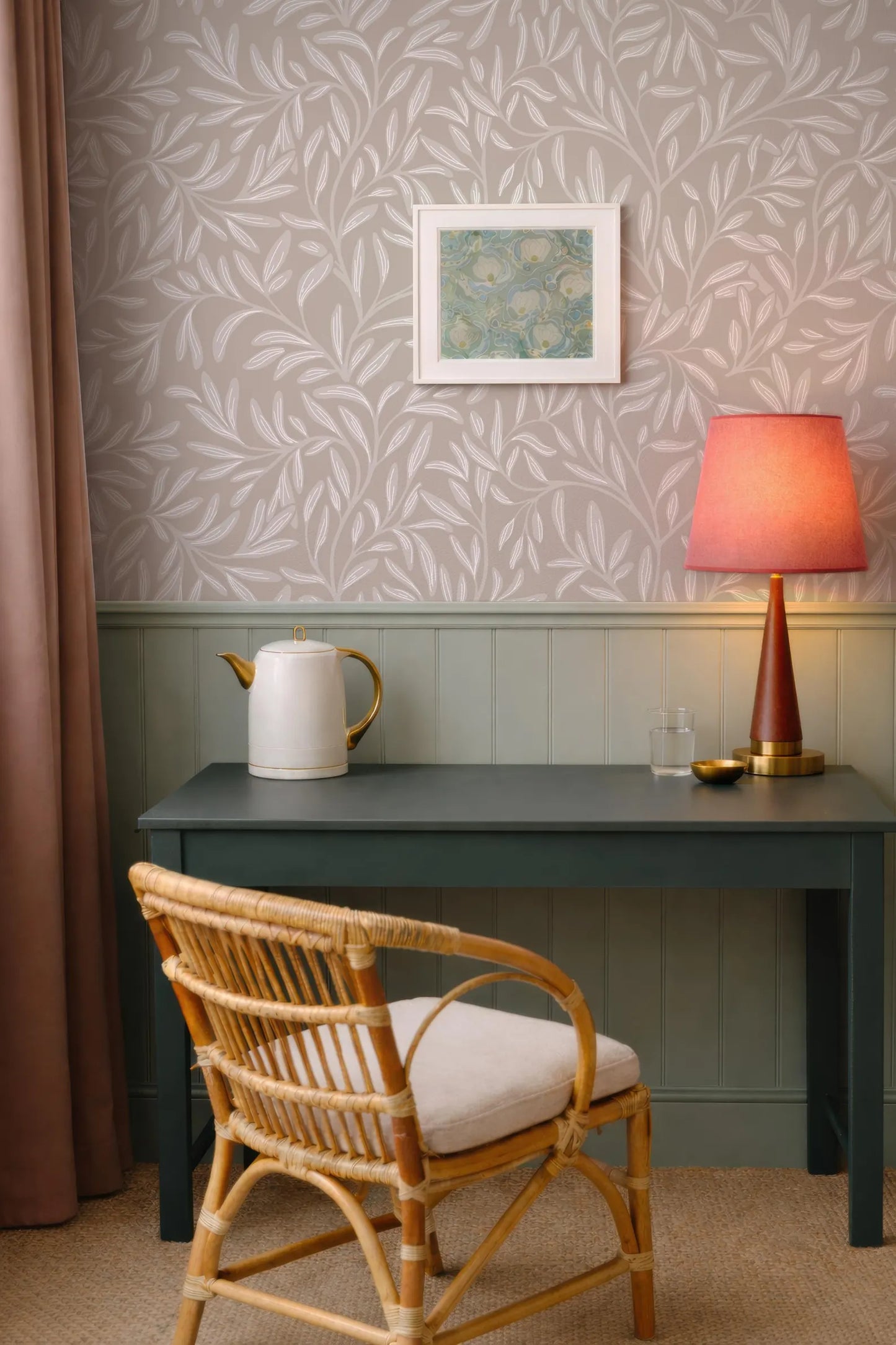 a cozy corner of a room with a green desk, a white teapot, a red lamp, and a wicker chair. The walls are adorned with a patterned wallpaper, and a framed picture is hanging above the desk.