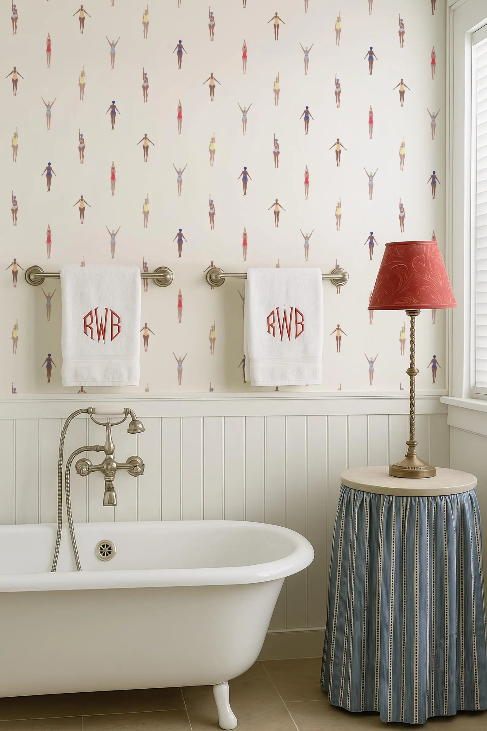 a bathroom with a clawfoot bathtub, two towels with initials, a red lamp, and a striped skirt.