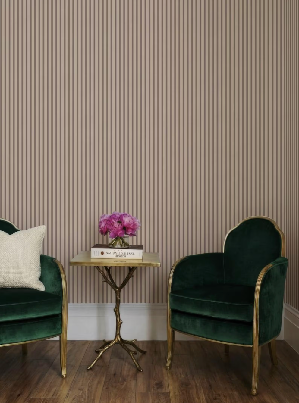 a cozy sitting area with two green velvet armchairs and a small side table with a vase of pink flowers. The wall behind the furniture has vertical stripes, creating a visually appealing backdrop.