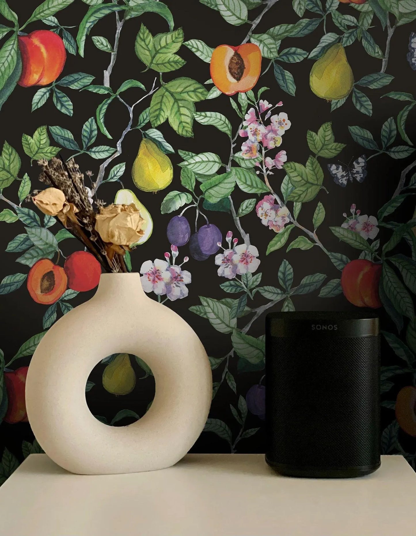 a white vase with a floral arrangement and a black speaker on a white surface, set against a black and green floral wallpaper.