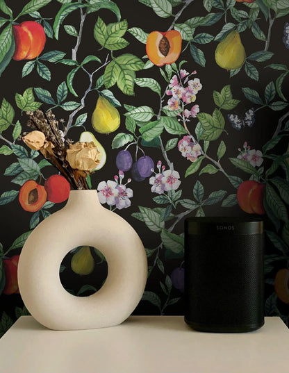 a white vase with a floral arrangement and a black speaker on a white surface, set against a black and green floral wallpaper.