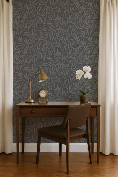 a wooden desk with a lamp, a clock, and a potted plant, situated in front of a gray wall with a leafy pattern.