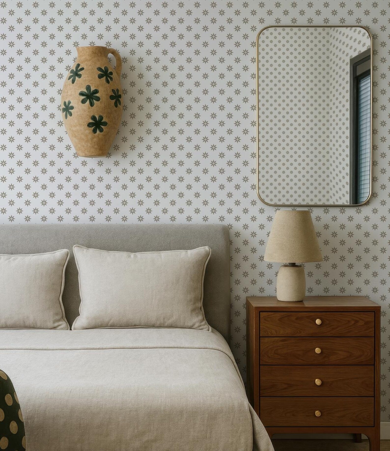 a bedroom with a bed, a mirror, and a wooden dresser. The bed has a gray headboard and white pillows. The mirror is mounted on the wall above the bed, and the dresser has a lamp on it. The wall has a patterned wallpaper with small white dots.