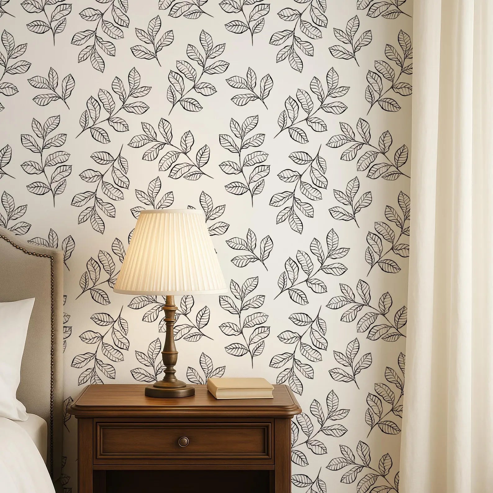 a bedroom setting with a wooden nightstand, a lamp, and a book on the nightstand. The wall behind the nightstand has a repeating pattern of black and white leaf designs.