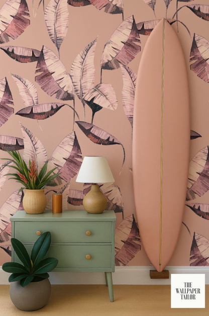 a vibrant pink wall with a large, colorful leaf pattern, a green dresser with a lamp, a potted plant, and a surfboard leaning against the wall.