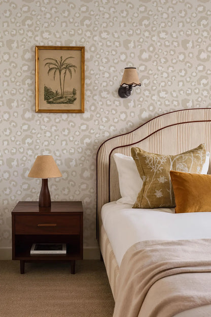 a cozy bedroom with a bed, a nightstand, and a framed picture on the wall. The bed has a striped headboard and is adorned with decorative pillows. The nightstand has a lamp on it, and there is a book on the nightstand as well. The wall behind the bed has a patterned wallpaper, and there is a picture hanging above the bed.