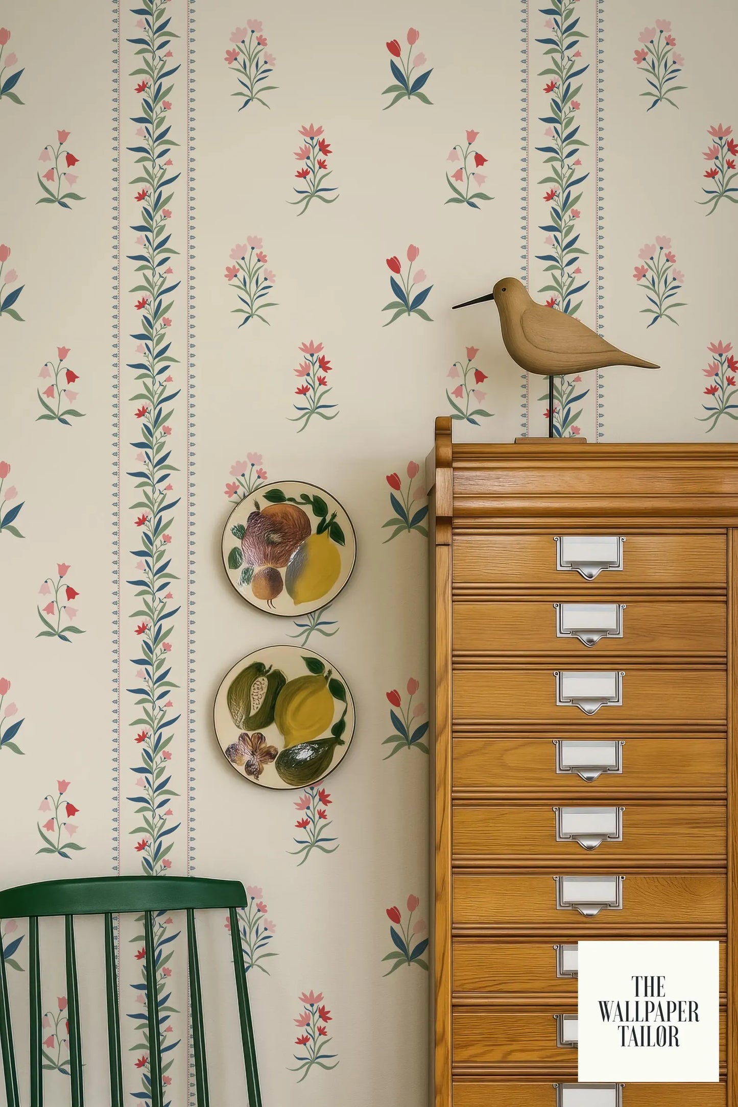 a wallpapered wall with a wooden cabinet, a green chair, and a bird figurine. There are two plates with fruit designs on the wall, and a bird figurine is perched on top of the cabinet.