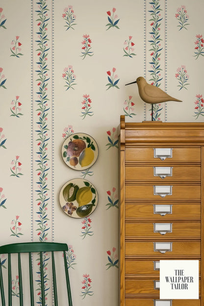 a wallpapered wall with a wooden cabinet, a green chair, and a bird figurine. There are two plates with fruit designs on the wall, and a bird figurine is perched on top of the cabinet.