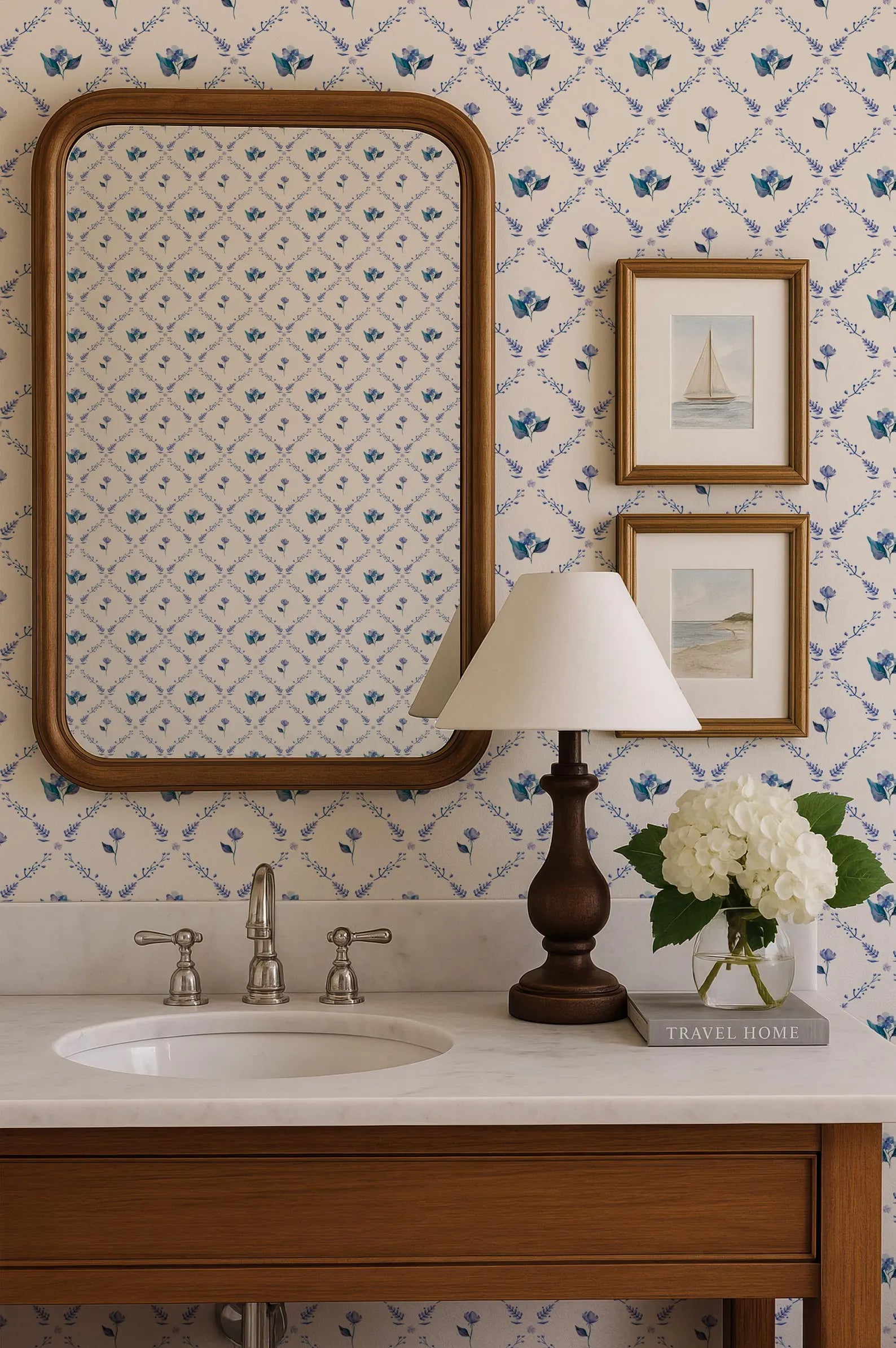 a bathroom vanity with a white sink, a mirror, a lamp, and a framed picture of a sailboat. The wall behind the vanity has a blue and white floral pattern.
