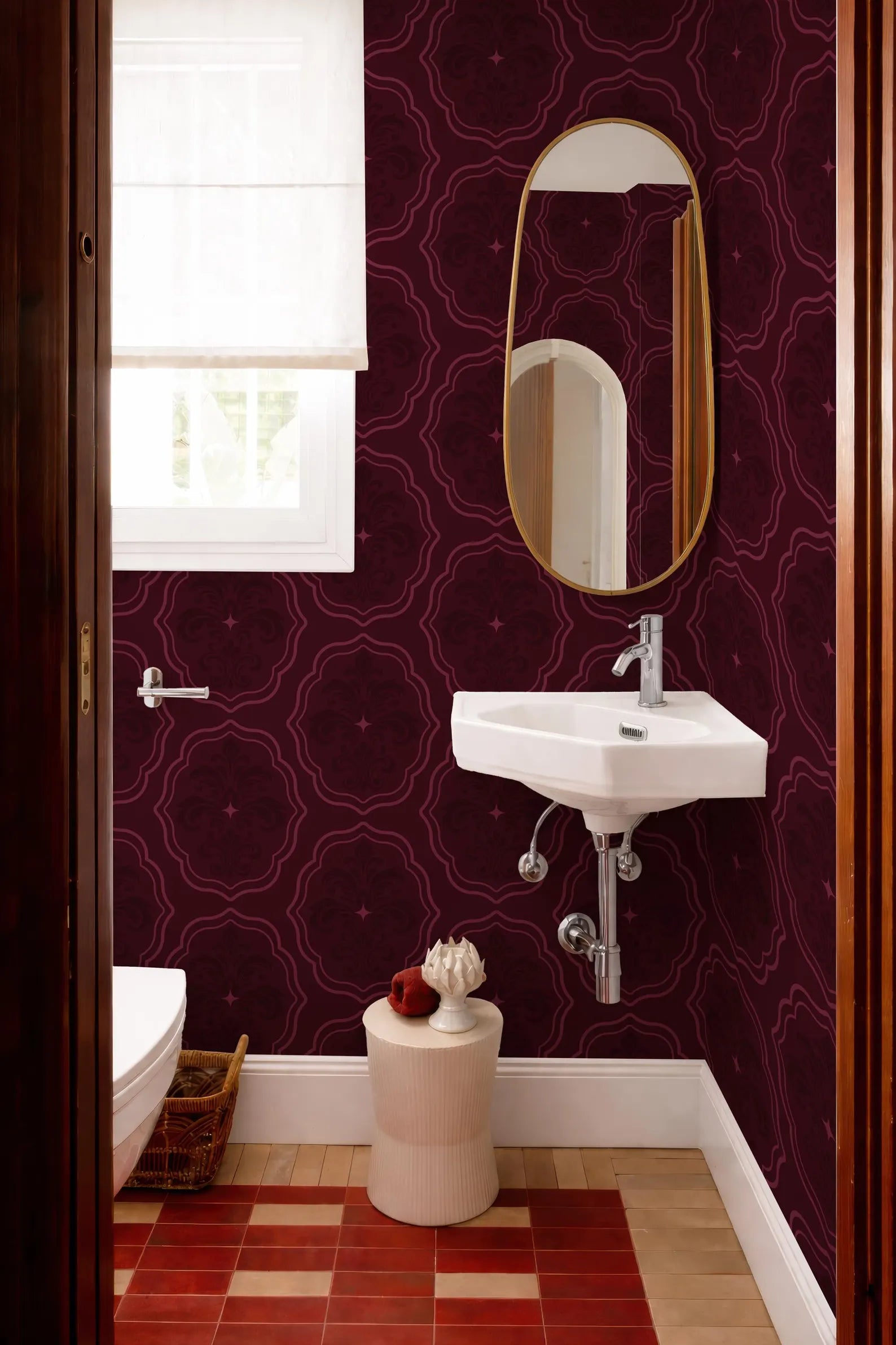 a bathroom with a white sink, a mirror, and a window. The walls are covered in a dark purple patterned wallpaper, and the floor is tiled in a red and white checkered pattern. There is a small white stool in front of the sink, and a basket is placed on the floor next to the toilet.