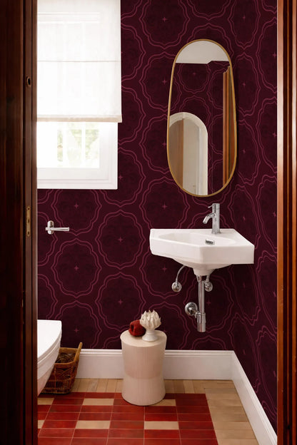 a bathroom with a white sink, a mirror, and a window. The walls are covered in a dark purple patterned wallpaper, and the floor is tiled in a red and white checkered pattern. There is a small white stool in front of the sink, and a basket is placed on the floor next to the toilet.