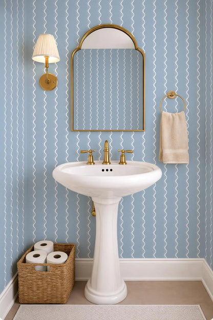 a bathroom with a white pedestal sink, a mirror, and a towel rack. The walls are covered in blue and white striped wallpaper, and there is a basket of toilet paper on the floor next to the sink.