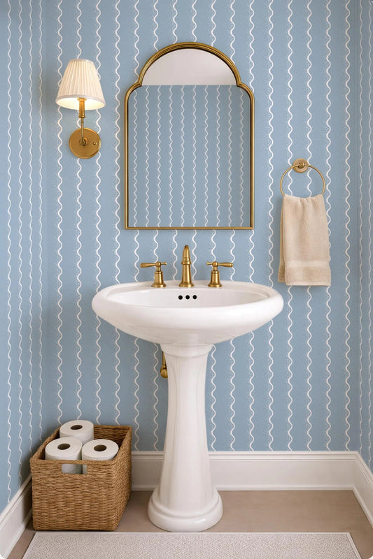 a bathroom with a white pedestal sink, a mirror, and a towel rack. The walls are covered in blue and white striped wallpaper, and there is a basket of toilet paper on the floor next to the sink.