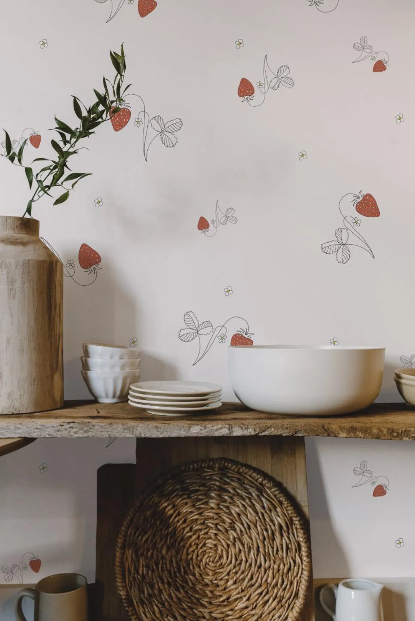 a shelf with various kitchen items, including a bowl, plates, a vase, and a woven basket. The wall behind the shelf has a repeating pattern of strawberries and leaves, creating a visually appealing and decorative backdrop.