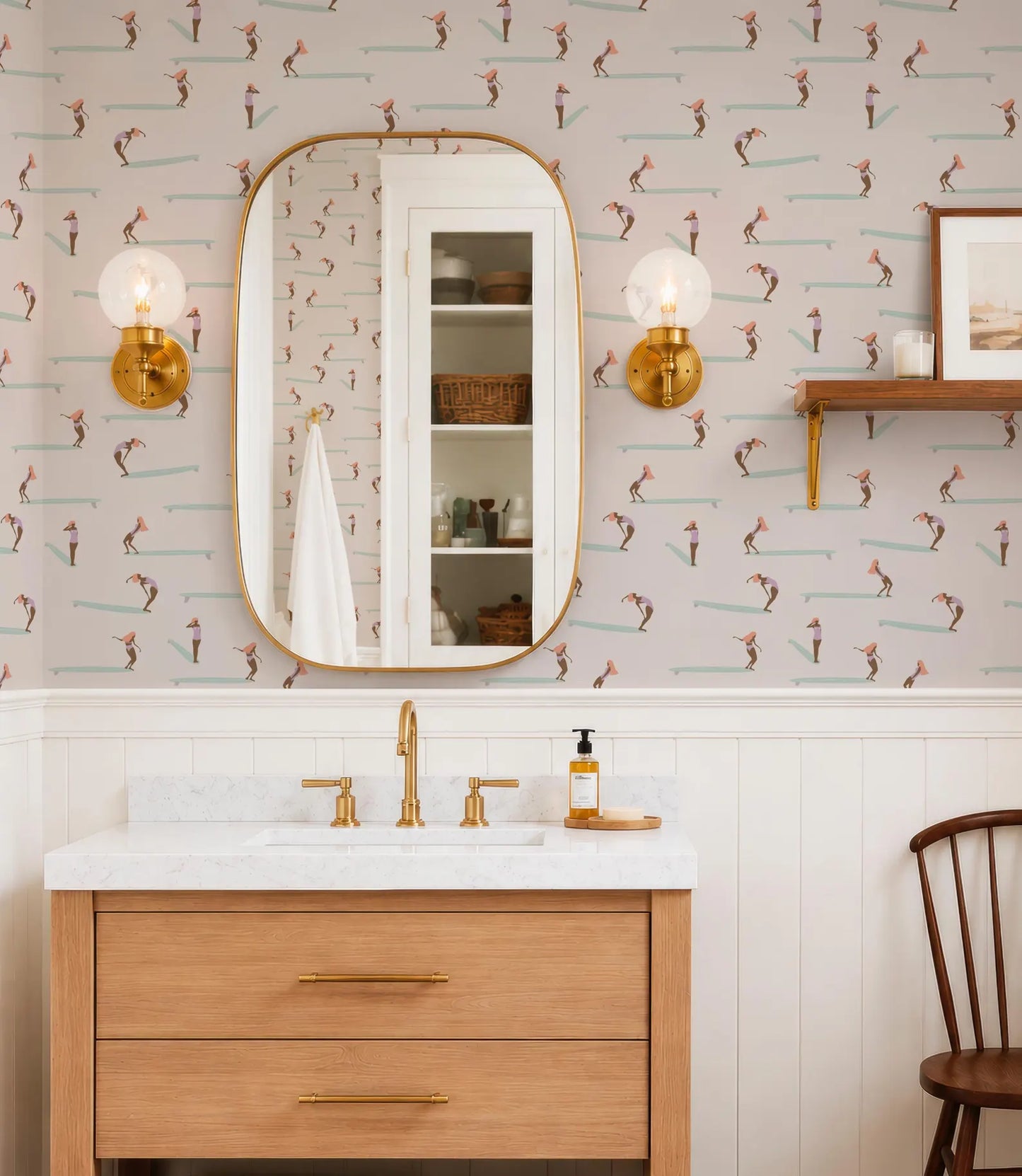 a bathroom with a white sink, a mirror, and a wooden vanity. The walls are decorated with a pattern of surfers on the wallpaper, and there are two gold-framed lights above the mirror.