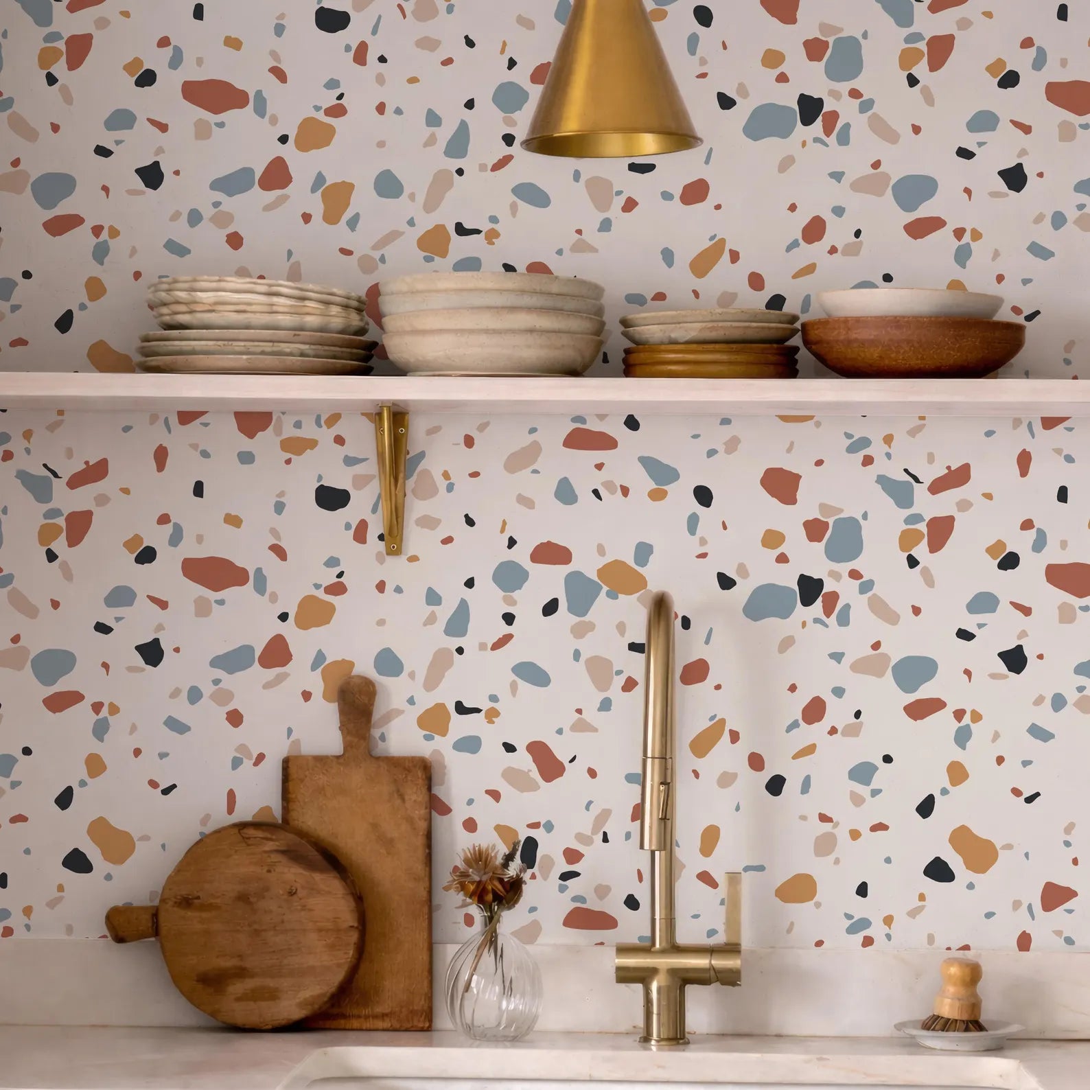 a kitchen counter with a variety of kitchen items, including wooden cutting boards, bowls, and a vase with flowers. The counter is set against a colorful, speckled wall with a gold pendant light hanging above it.