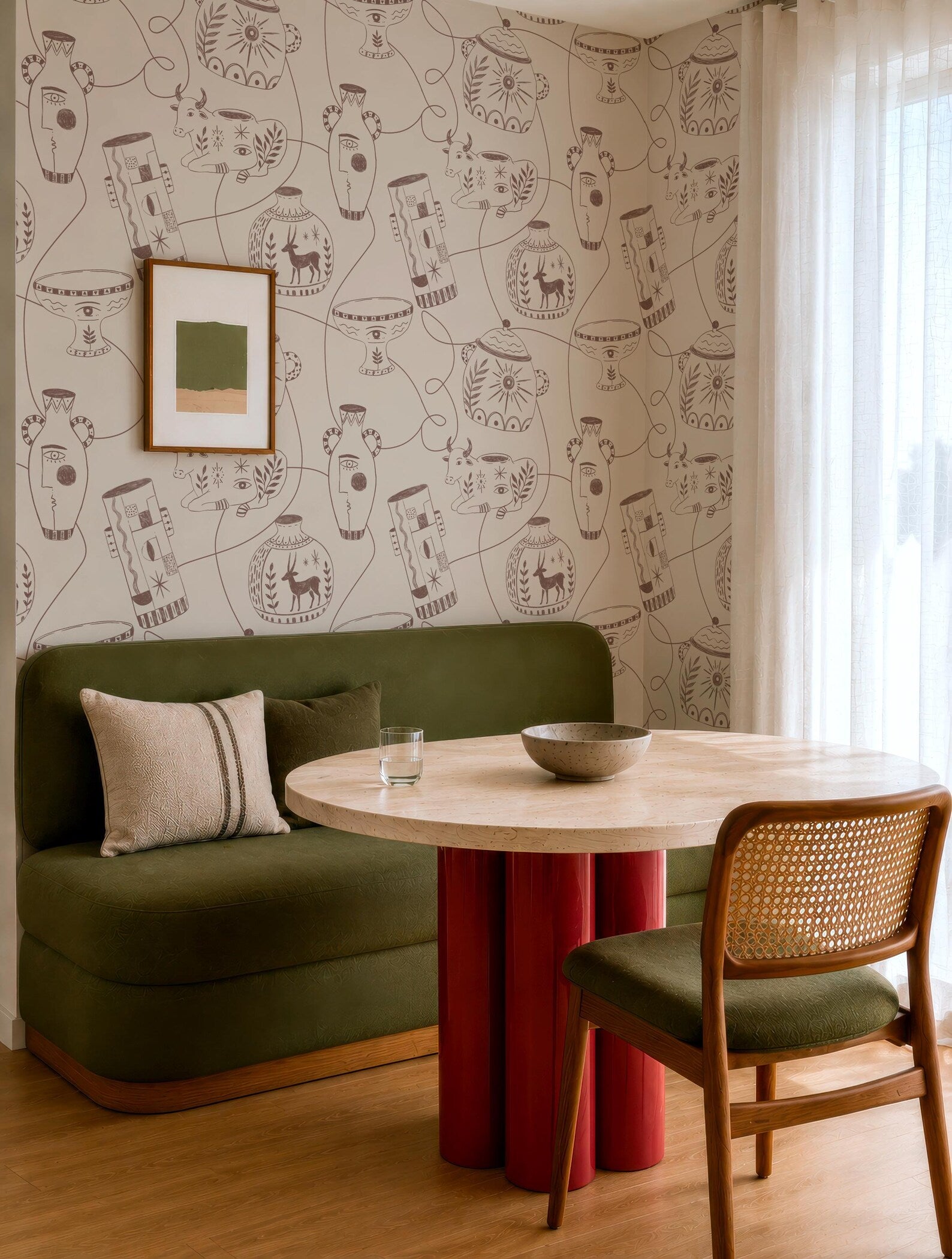 a cozy living room with a green sofa, a round wooden table with a bowl on it, and a woven chair. The walls are decorated with a patterned wallpaper, and there is a framed picture hanging on the wall.