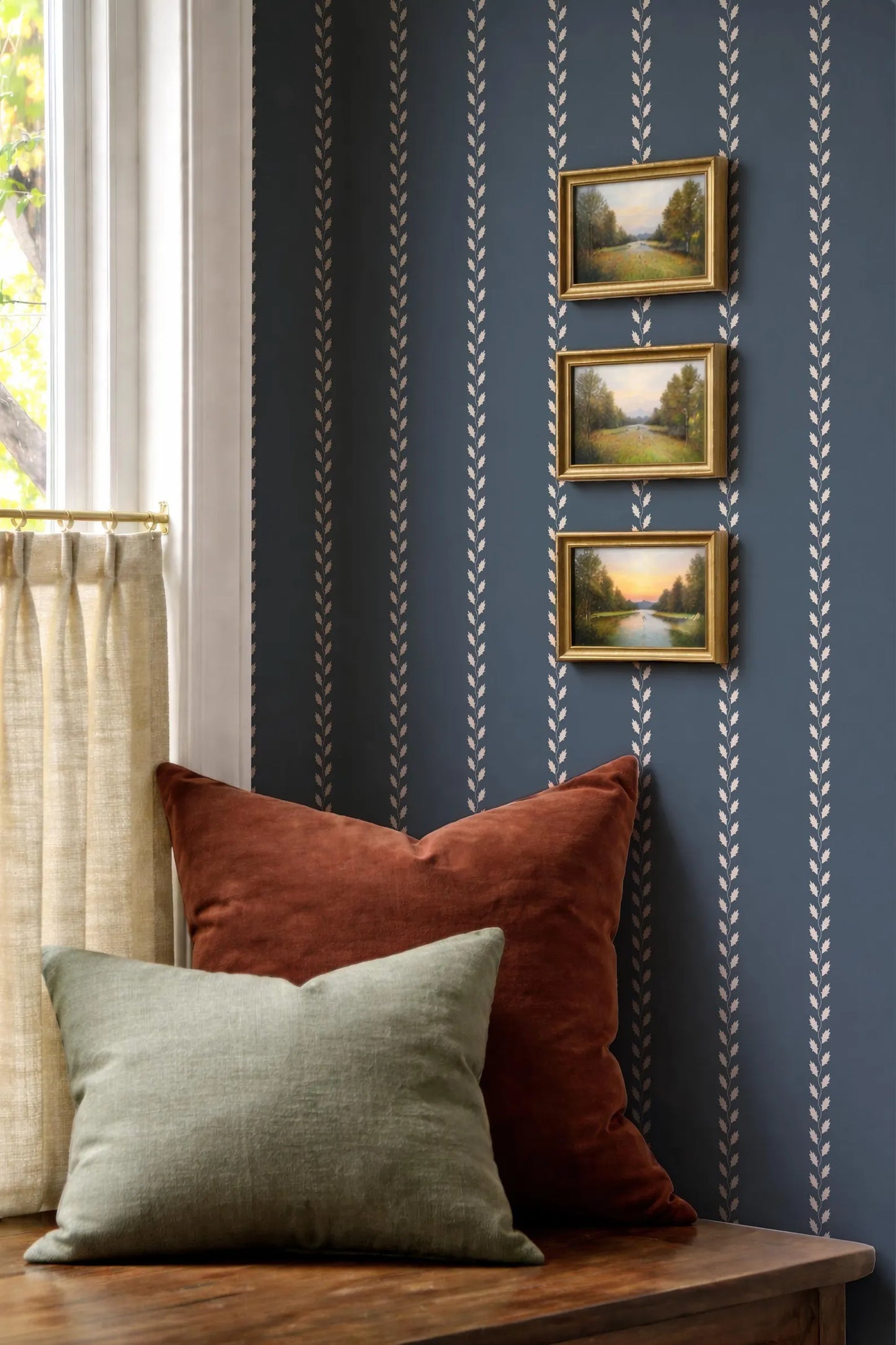 a cozy corner with a wooden table, a window with white curtains, and three framed landscape paintings on the wall.
