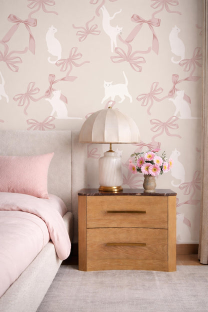 a bedroom scene with a bed, a wooden nightstand, and a lamp with a white shade. The wall behind the bed has a pink and white cat wallpaper pattern.