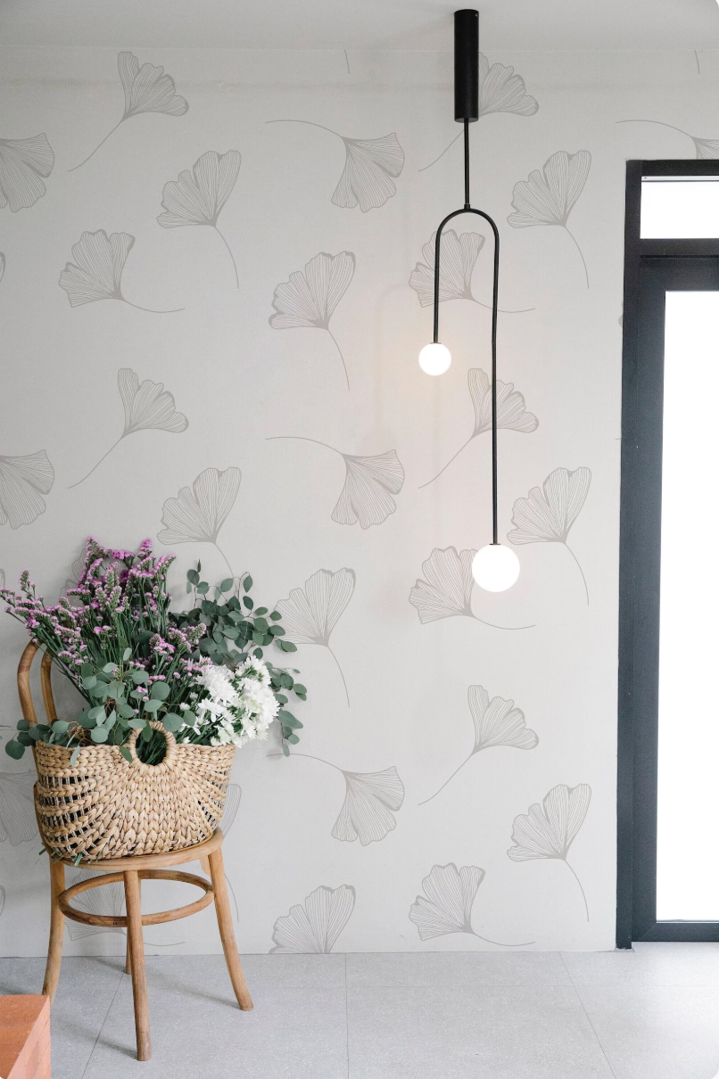 a woven basket filled with flowers and greenery, placed on a wooden chair against a wall with a repeating pattern of black and white gilt leaves. A hanging light fixture with two round white globes is visible in the background.