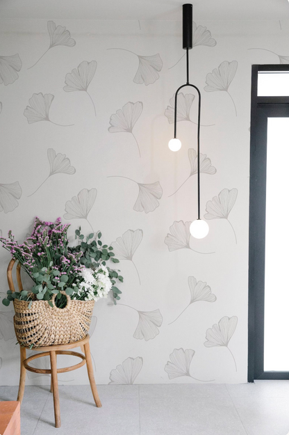 a woven basket filled with flowers and greenery, placed on a wooden chair against a wall with a repeating pattern of black and white gilt leaves. A hanging light fixture with two round white globes is visible in the background.
